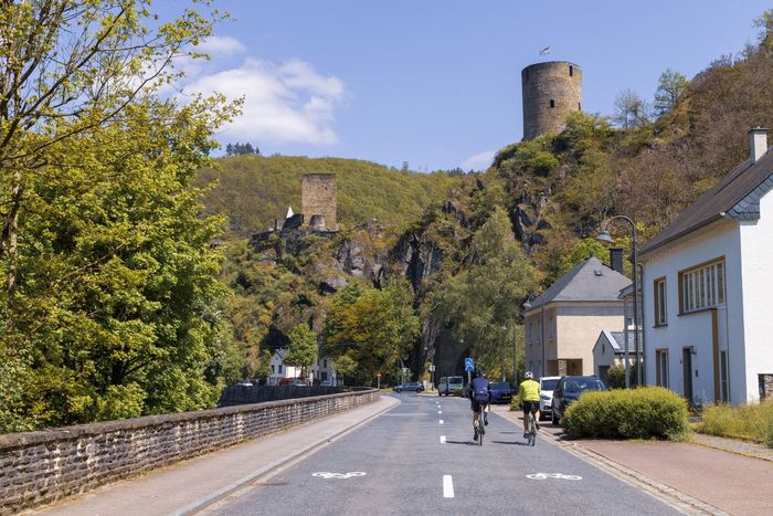 Two cyclists ride side by side on a road in Esch-sur-Sûre. A part of Esch-sur-Sûre Castle is visible in the background.