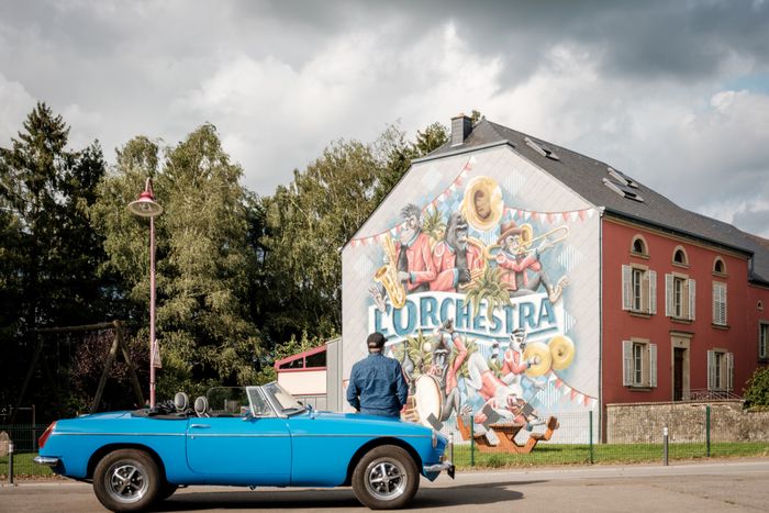 A man admires a mural painting while taking a break, leaning on the hood of his blue classic car