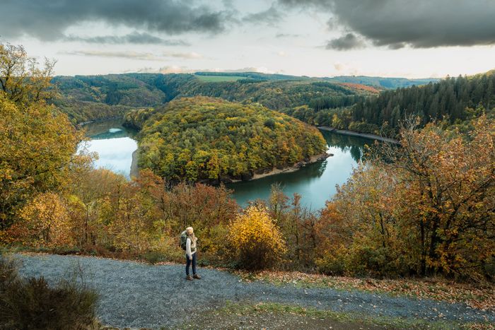 Eine Frau spaziert auf einem Kiesweg entlang des Éislek Pad Bavigne. Sie befindet sich am Aussichtspunkt Schäsbierg und genießt den atemberaubenden Blick auf die herbstliche Flussschleife.